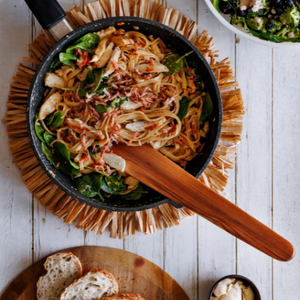 A close-up, action-oriented product photograph of a handcrafted 37cm teak wood sauté spatula lifting chicken and fettuccine pasta out of a hot black cast iron skillet. The flat, straight edge of the blade is shown under the food, showcasing its structural strength. In the background, a rustic table setting features a fresh green salad in a large bowl and sliced sourdough bread on a wooden board. The prominent, swirling grain of the single-piece sustainable teak tool stands out vividly.