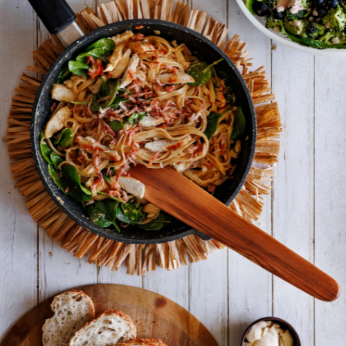 A close-up, action-oriented product photograph of a handcrafted 37cm teak wood sauté spatula lifting chicken and fettuccine pasta out of a hot black cast iron skillet. The flat, straight edge of the blade is shown under the food, showcasing its structural strength. In the background, a rustic table setting features a fresh green salad in a large bowl and sliced sourdough bread on a wooden board. The prominent, swirling grain of the single-piece sustainable teak tool stands out vividly.