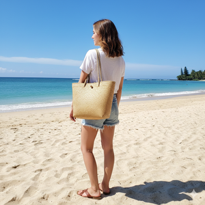 A woman in denim shorts and a white t-shirt on a sandy beach, carrying a large hand-woven natural sea grass tote bag with matching rope handles on her shoulder, demonstrating its size as a perfect beach bag.