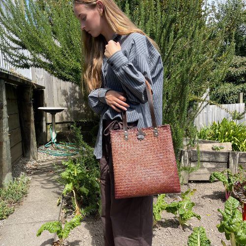 A woman wearing a striped shirt and dark trousers carrying a mahogany brown hand-woven Krajood sea grass bag, demonstrating its size and style in a sunny outdoor garden setting.