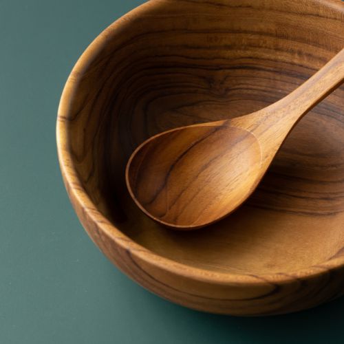 A detailed close-up of a handcrafted teak wood soup spoon resting inside a solid teak bowl, highlighting the intricate, swirling natural wood grain and the smooth, food-safe finish of the artisan-made dinnerware set.