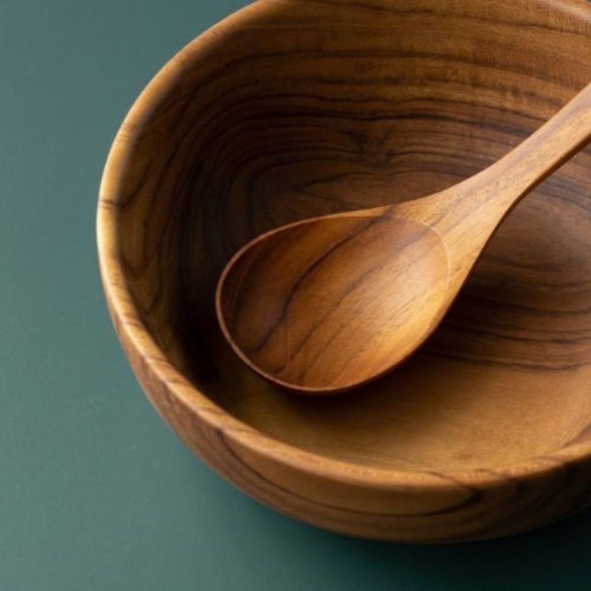 A detailed close-up of a handcrafted teak wood soup spoon resting inside a solid teak bowl, highlighting the intricate, swirling natural wood grain and the smooth, food-safe finish of the artisan-made dinnerware set.