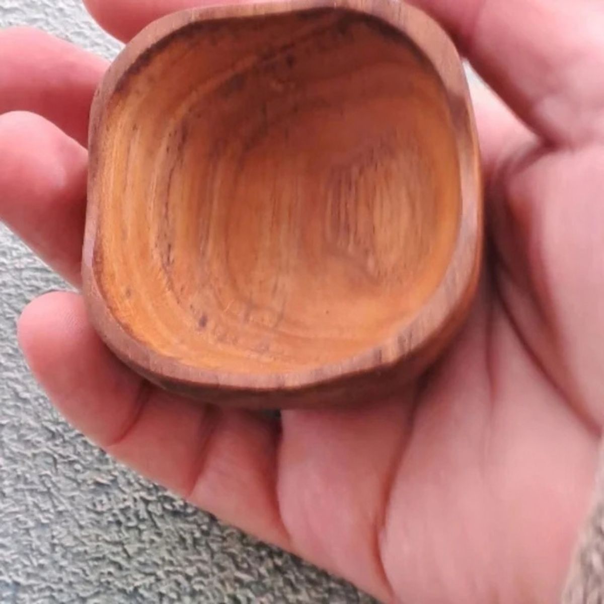 A close-up lifestyle photograph showing a small, square handcrafted pinch bowl carved from sustainable teak wood resting in the open palm of a person's hand. The overhead perspective clearly demonstrates the compact size and scale of the dish, highlighting its softly rounded square rim, deep smooth interior, and rich golden-brown timber tones with a prominent natural wood grain. Carved from a single piece of non-toxic teak, this durable mini pot is designed for holding spices, salts, or condiments.