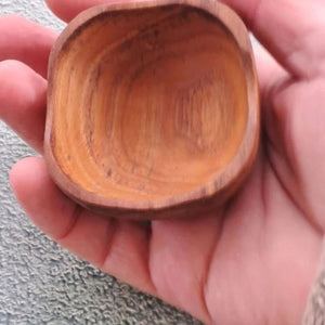 A close-up lifestyle photograph showing a small, square handcrafted pinch bowl carved from sustainable teak wood resting in the open palm of a person's hand. The overhead perspective clearly demonstrates the compact size and scale of the dish, highlighting its softly rounded square rim, deep smooth interior, and rich golden-brown timber tones with a prominent natural wood grain. Carved from a single piece of non-toxic teak, this durable mini pot is designed for holding spices, salts, or condiments.