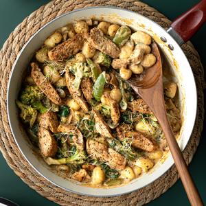 A long-handled teak wooden spoon resting inside a deep ceramic frying pan filled with a hot, creamy chicken and gnocchi stir-fry with broccoli. The image showcases the spoon's extra-long handle extending well past the rim of the pan, demonstrating its practical utility for deep-pan cooking, sautéing, and keeping hands safely away from heat.