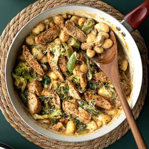 A long-handled teak wooden spoon resting inside a deep ceramic frying pan filled with a hot, creamy chicken and gnocchi stir-fry with broccoli. The image showcases the spoon's extra-long handle extending well past the rim of the pan, demonstrating its practical utility for deep-pan cooking, sautéing, and keeping hands safely away from heat.