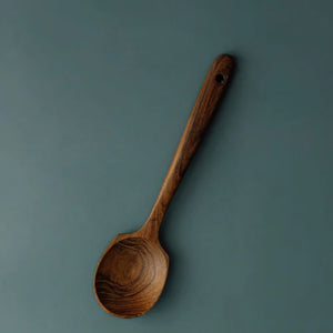 A handcrafted teak wood spoon with a uniquely wide, shallow head and a long ergonomic handle, featuring a rich dark brown grain and a smooth finish, isolated against a muted teal-grey background.