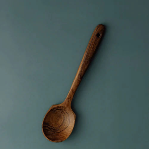 A handcrafted teak wood spoon with a uniquely wide, shallow head and a long ergonomic handle, featuring a rich dark brown grain and a smooth finish, isolated against a muted teal-grey background.