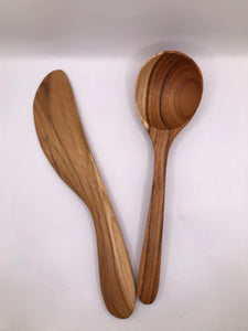 A pair of handcrafted teak wood kitchen utensils, including a large serving spoon and a curved spread knife, showcasing matching golden-brown wood grains and a smooth, non-toxic finish against a white background.