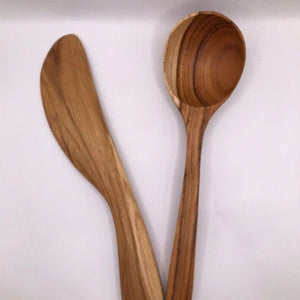 A pair of handcrafted teak wood kitchen utensils, including a large serving spoon and a curved spread knife, showcasing matching golden-brown wood grains and a smooth, non-toxic finish against a white background.