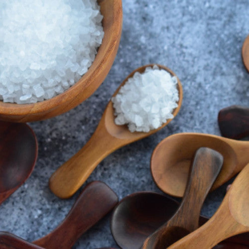 A close-up view of multiple handcrafted teak wood salt spoons in both light and dark mahogany-toned wood grains, with one spoon prominently displaying coarse white sea salt next to a matching wooden pinch bowl.