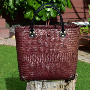 A hand-woven Krajood sea grass tote bag in a deep burgundy hue, featuring rolled black leather handles secured with brass-toned rivets and reinforced leather corner protectors, resting on a rustic wooden stump.