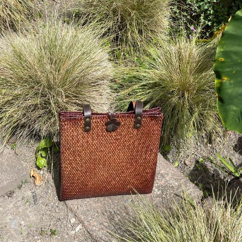 A hand-woven Krajood sea grass shoulder bag in a rich mahogany brown, featuring durable leather strap handles with brass-toned rivets and a decorative leather flower clasp, displayed in a natural outdoor garden setting.