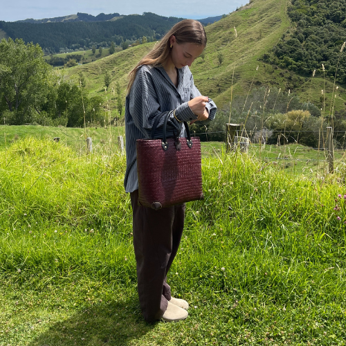 A hand-woven Krajood sea grass shoulder bag in a rich, uniform mahogany brown, featuring dark leather straps with brass-toned rivets and a handcrafted leather flower clasp, displayed in a natural garden setting.