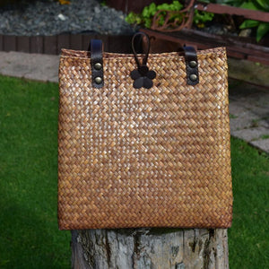 A hand-woven mahogany brown Krajood sea grass bag displayed outdoors on a natural wood tree stump, showing the tight artisan weave, dark leather handles, and a decorative flower clasp.