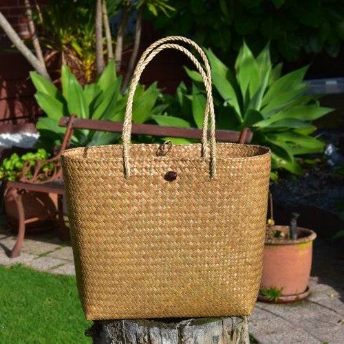 A hand-woven natural Krajood sea grass tote bag featuring matching woven rope-style handles and a dark wooden bead toggle closure, displayed outdoors on a tree stump.