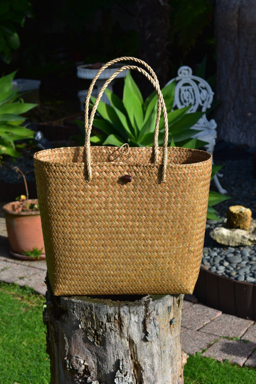 A hand-woven natural Krajood sea grass tote bag featuring matching woven rope-style handles and a dark wooden bead toggle closure, displayed outdoors on a tree stump.