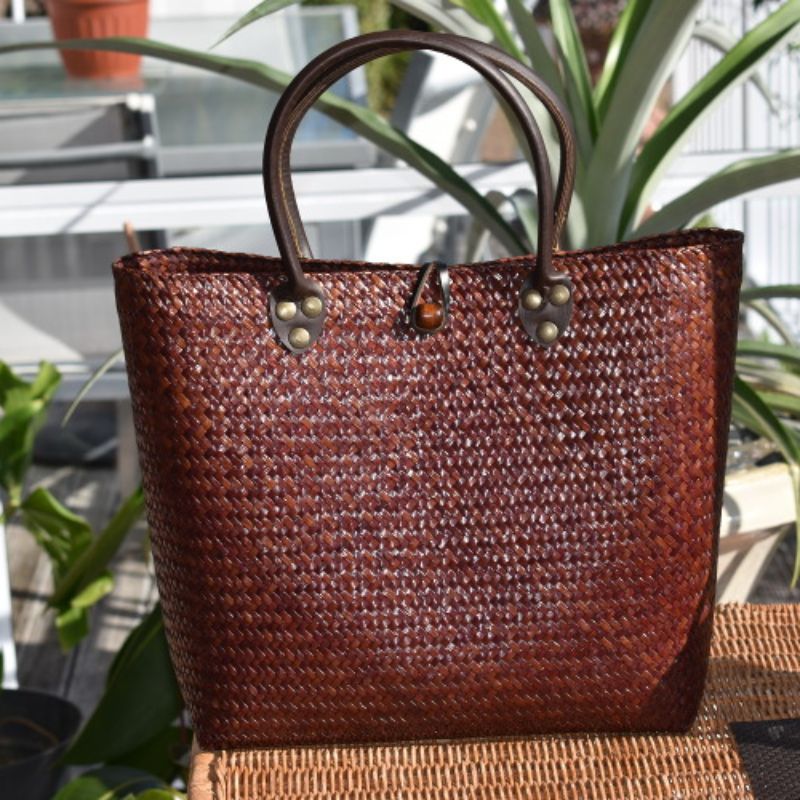 A close-up of a hand-woven Krajood sea grass basket in a deep mahogany brown, featuring a sophisticated woven pattern and durable dark leather handles secured with brass-toned rivets.