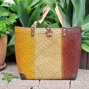 A hand-woven Krajood sea grass tote bag featuring a vertical three-tone gradient of natural, tan, and dark brown, finished with genuine leather strap handles and a flower-shaped clasp.