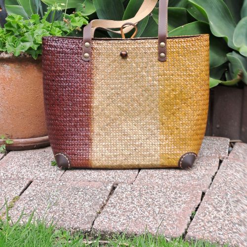 An artisan-crafted Krajood sea grass bag featuring honey, tan, and burgundy vertical panels, displayed outdoors on a stone patio with lush green foliage.