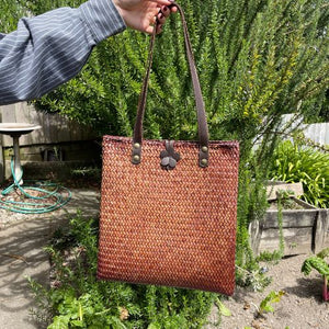 A hand-woven Krajood sea grass shoulder bag in rich mahogany brown, held by its long leather straps to show the bag's height and the decorative leather flower clasp in a sunny garden setting.