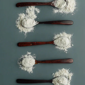 A top-down artistic arrangement of five handcrafted rosewood dessert spoons on a muted teal background, each cradling a dusting of white flour to highlight the deep, dark wood grain and smooth polished surface