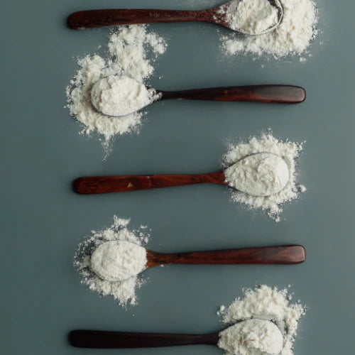 A top-down artistic arrangement of five handcrafted rosewood dessert spoons on a muted teal background, each cradling a dusting of white flour to highlight the deep, dark wood grain and smooth polished surface
