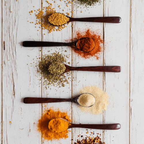 A top-down artistic shot of five dark rosewood teaspoons arranged on a rustic white wood surface, each holding a different colorful spice including turmeric, paprika, and dried herbs, showcasing the rich wood grain against the vibrant textures  part of a 6 piece set