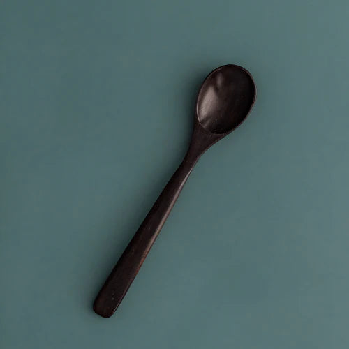 A single handcrafted rosewood teaspoon featuring a rich, dark espresso-toned finish and an elegant tapered handle, angled against a muted teal-grey background to showcase its smooth organic texture and artisanal quality