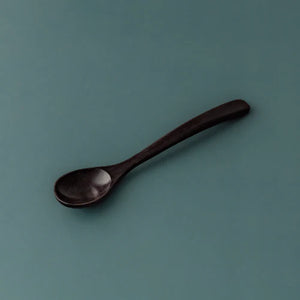 A single handcrafted rosewood teaspoon with a dark, rich espresso-toned finish and an elegant tapered handle, angled against a muted teal-grey background to highlight its smooth organic texture.