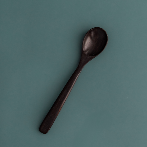 A single handcrafted rosewood teaspoon with a dark, rich espresso-toned finish and an elegant tapered handle, angled against a muted teal-grey background to highlight its smooth organic texture.