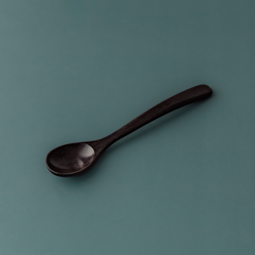 A single handcrafted rosewood teaspoon with a dark, rich espresso-toned finish and an elegant tapered handle, angled against a muted teal-grey background to highlight its smooth organic texture.  Part of a 6 piece set.