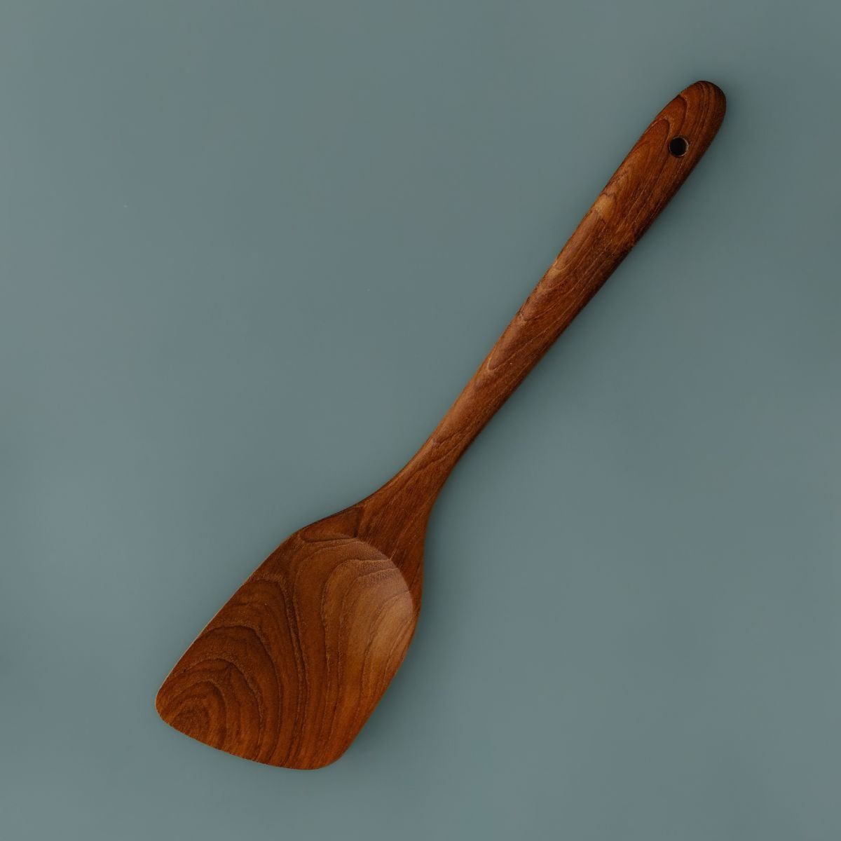 Single handcrafted teak wooden spatula with a rich natural grain finish and angled blade, lying on a smooth slate blue background.