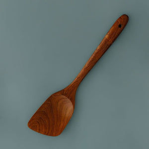Single handcrafted teak wooden spatula with a rich natural grain finish and angled blade, lying on a smooth slate blue background.