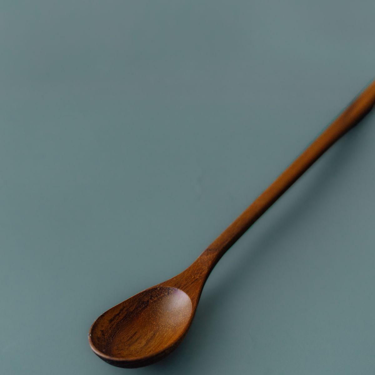Angled close-up of Yompai's handcrafted wooden parfait spoon, showing the shallow oval bowl and slim teak handle on a muted blue background.