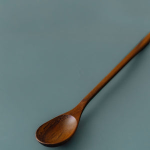Angled close-up of Yompai's handcrafted wooden parfait spoon, showing the shallow oval bowl and slim teak handle on a muted blue background.