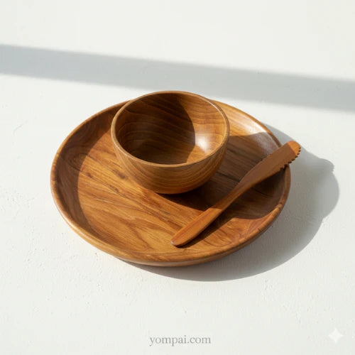 A top-down lifestyle shot of a sustainable teak wood entertaining set, including a large round platter, a matching small bowl, and a serrated wooden knife, showcasing the rich golden-brown wood grain under bright, natural sunlight on a white surface