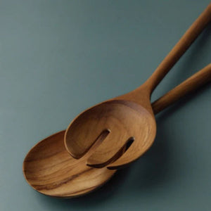 An angled, close-up view of a large teak wood salad server set, showing a solid spoon and a three-tine server fork. The image highlights the natural, swirling wood grain and the smooth, ergonomic long handles against a soft teal background.