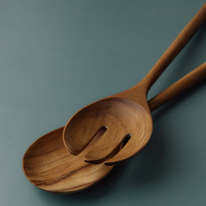An angled, close-up view of a large teak wood salad server set, showing a solid spoon and a three-tine server fork. The image highlights the natural, swirling wood grain and the smooth, ergonomic long handles against a soft teal background.