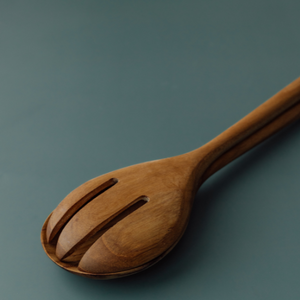 A close-up, angled view of a handcrafted teak wood salad server set, showing the three-tine server fork nested perfectly on top of the solid salad spoon. The image highlights the smooth, carved edges and the rich, natural wood grain against a matte teal background.