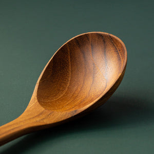 An extreme close-up of the rounded bowl of a handcrafted teak wood soup spoon. The image highlights the intricate, shimmering grain patterns and the smooth, carved edges of the teak, set against a matte dark green background.