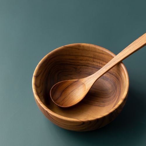 A top-down product shot of a handcrafted teak wood soup spoon resting inside a matching solid teak bowl, set against a smooth sage green background to highlight the intricate natural wood grain and golden-brown tones.
