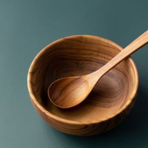 A top-down product shot of a handcrafted teak wood soup spoon resting inside a matching solid teak bowl, set against a smooth sage green background to highlight the intricate natural wood grain and golden-brown tones.
