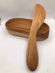 A handcrafted teak wood spread knife resting across a unique, oblong-curved wooden serving bowl, showcasing matching golden-brown wood grains and a smooth, non-toxic finish for an elegant tabletop presentation.