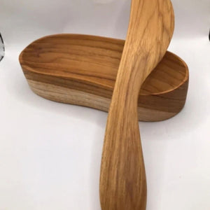 A handcrafted teak wood spread knife resting across a unique, oblong-curved wooden serving bowl, showcasing matching golden-brown wood grains and a smooth, non-toxic finish for an elegant tabletop presentation.
