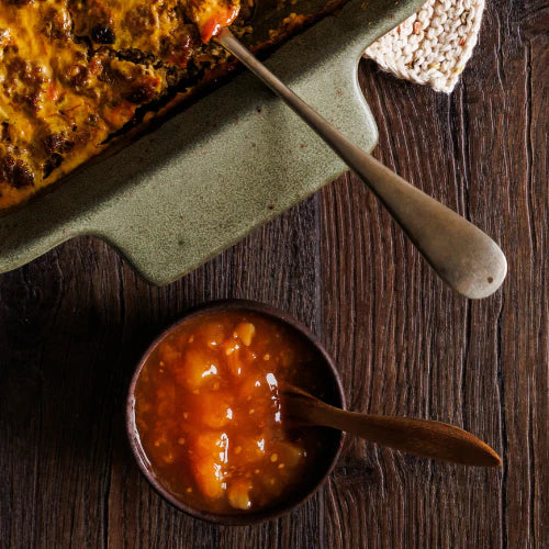 A set of handcrafted teak wood teaspoons with unique angled handles, featured in a lifestyle setting alongside a bowl of fruit preserves and a warm baked dish, showcasing the rich natural wood grain on a rustic dark wood surface.