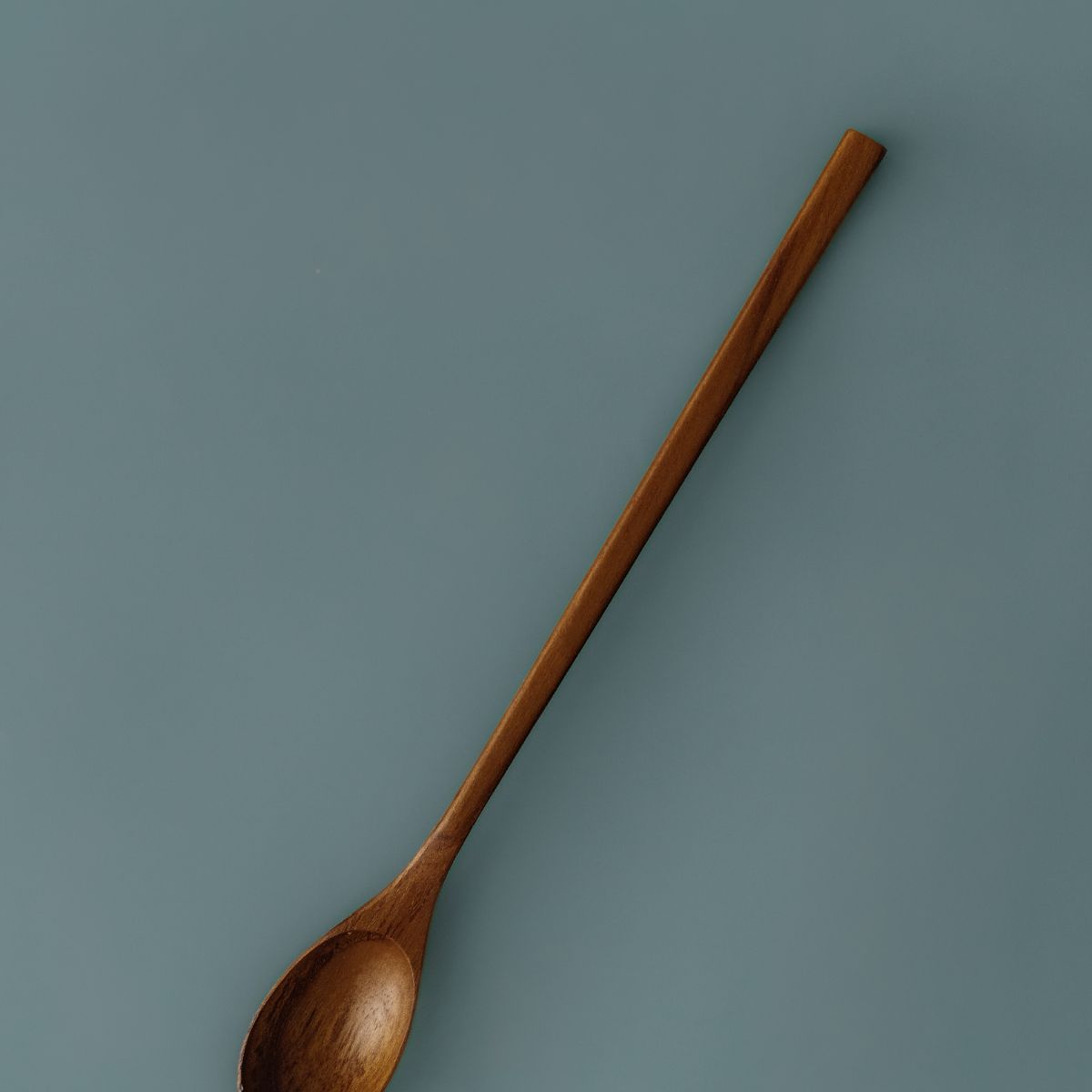 A professional product photograph of a handcrafted teak wood parfait spoon displayed diagonally against a teal background. The spoon features an extra-long, slender handle and a smooth, oval-shaped head, expertly carved from a single piece of sustainable teak. The image highlights the rich, warm natural grain and food-safe finish of the wood, emphasizing its durable and non-toxic design for reaching into tall jars, stirring lattes, or serving desserts.