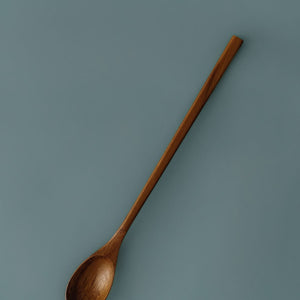 A professional product photograph of a handcrafted teak wood parfait spoon displayed diagonally against a teal background. The spoon features an extra-long, slender handle and a smooth, oval-shaped head, expertly carved from a single piece of sustainable teak. The image highlights the rich, warm natural grain and food-safe finish of the wood, emphasizing its durable and non-toxic design for reaching into tall jars, stirring lattes, or serving desserts.