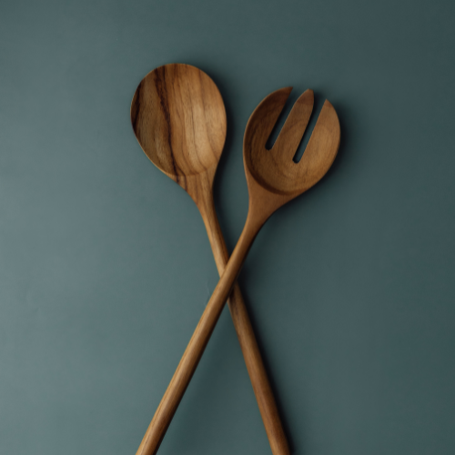 A top-down view of a large teak wood salad server set with the long handles crossed, displaying a solid spoon and a three-tine server fork. The image highlights the distinctive dark and light wood grain patterns and smooth finish against a matte slate blue background.