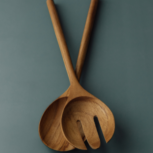 A close-up, top-down view of a handcrafted large teak wood salad server set, featuring one solid spoon and one pronged server with a three-tine detail. The set shows a rich, natural golden-brown grain and smooth finish against a matte slate blue background.